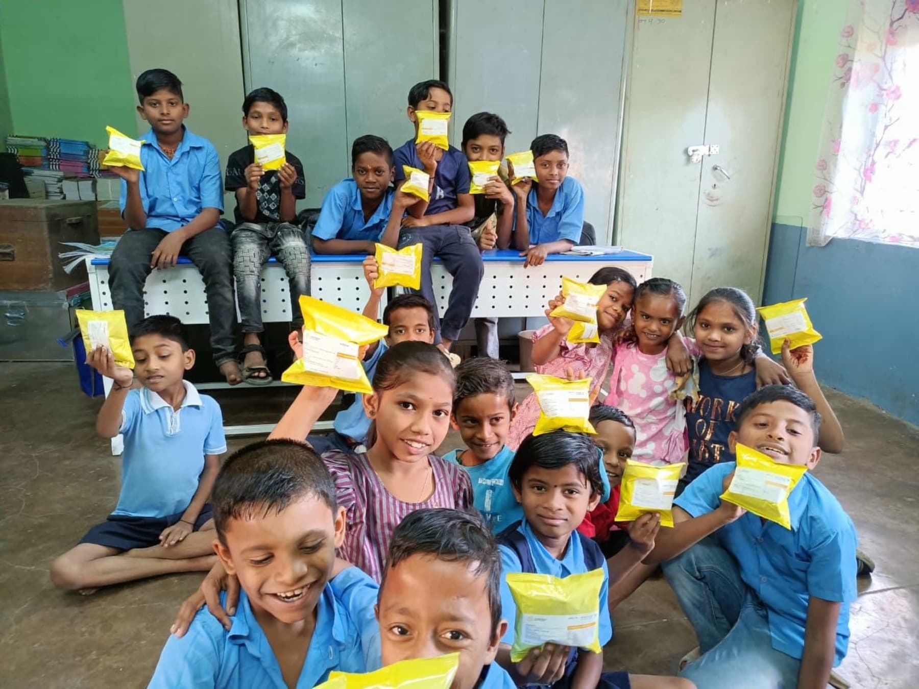 A classroom of children in blue uniforms hold up their yellow nutrition sachets, smiling.