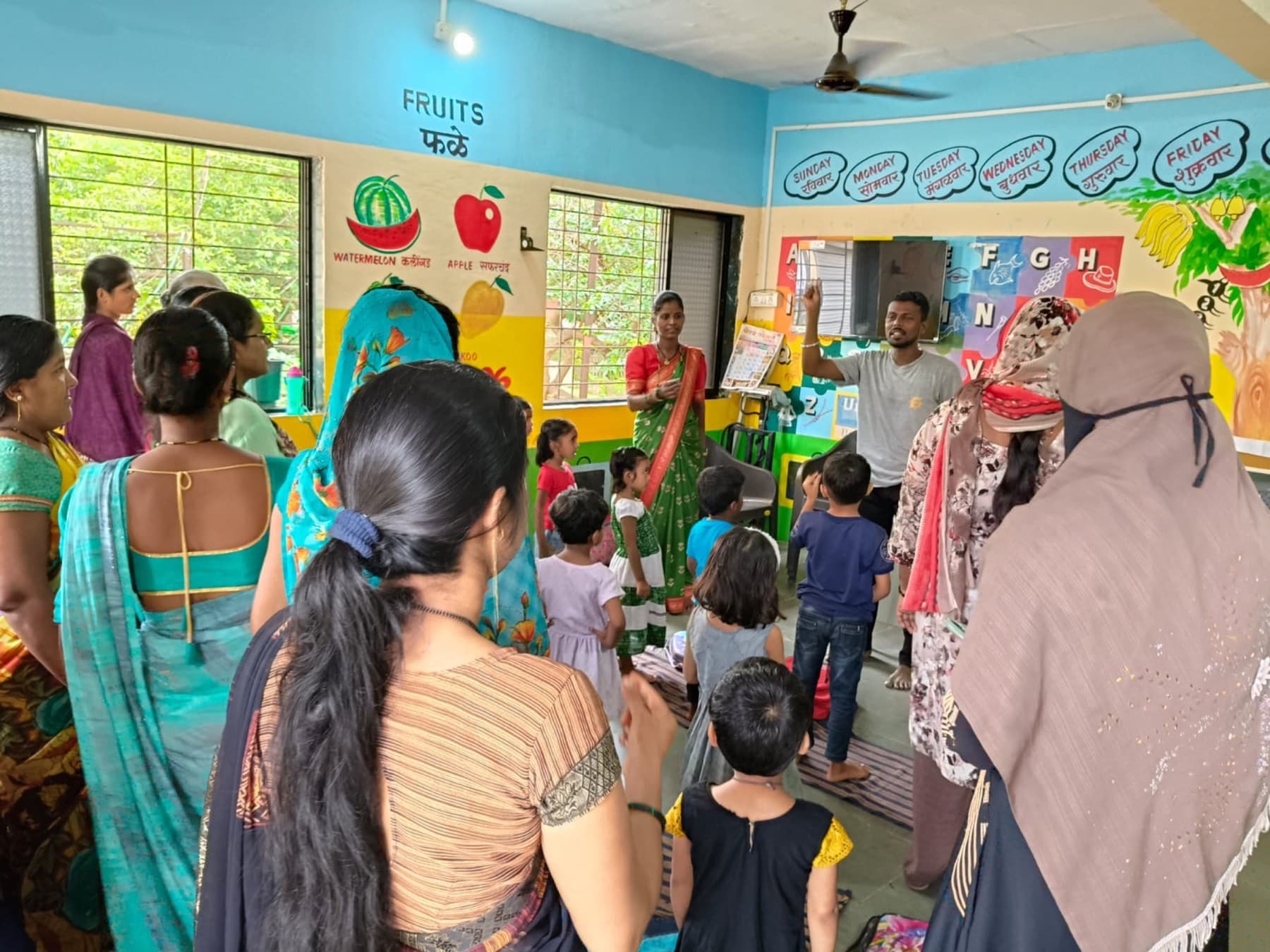 Women and young children stand together in a brightly painted anganwadi room while a facilitator leads a session.