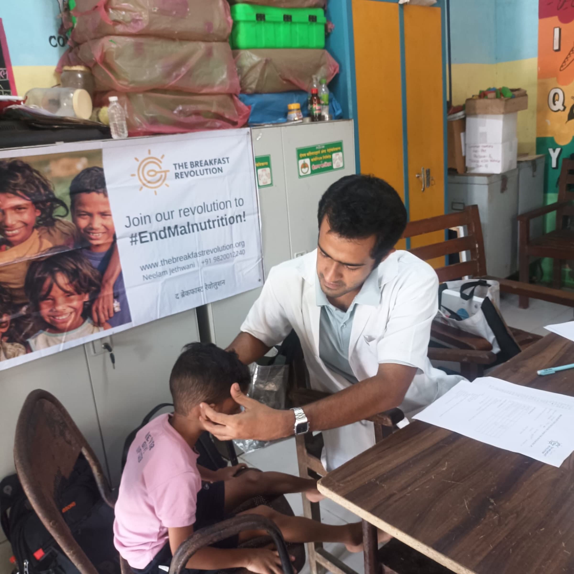 A field worker examines a child at a screening desk beneath a Breakfast Revolution banner.
