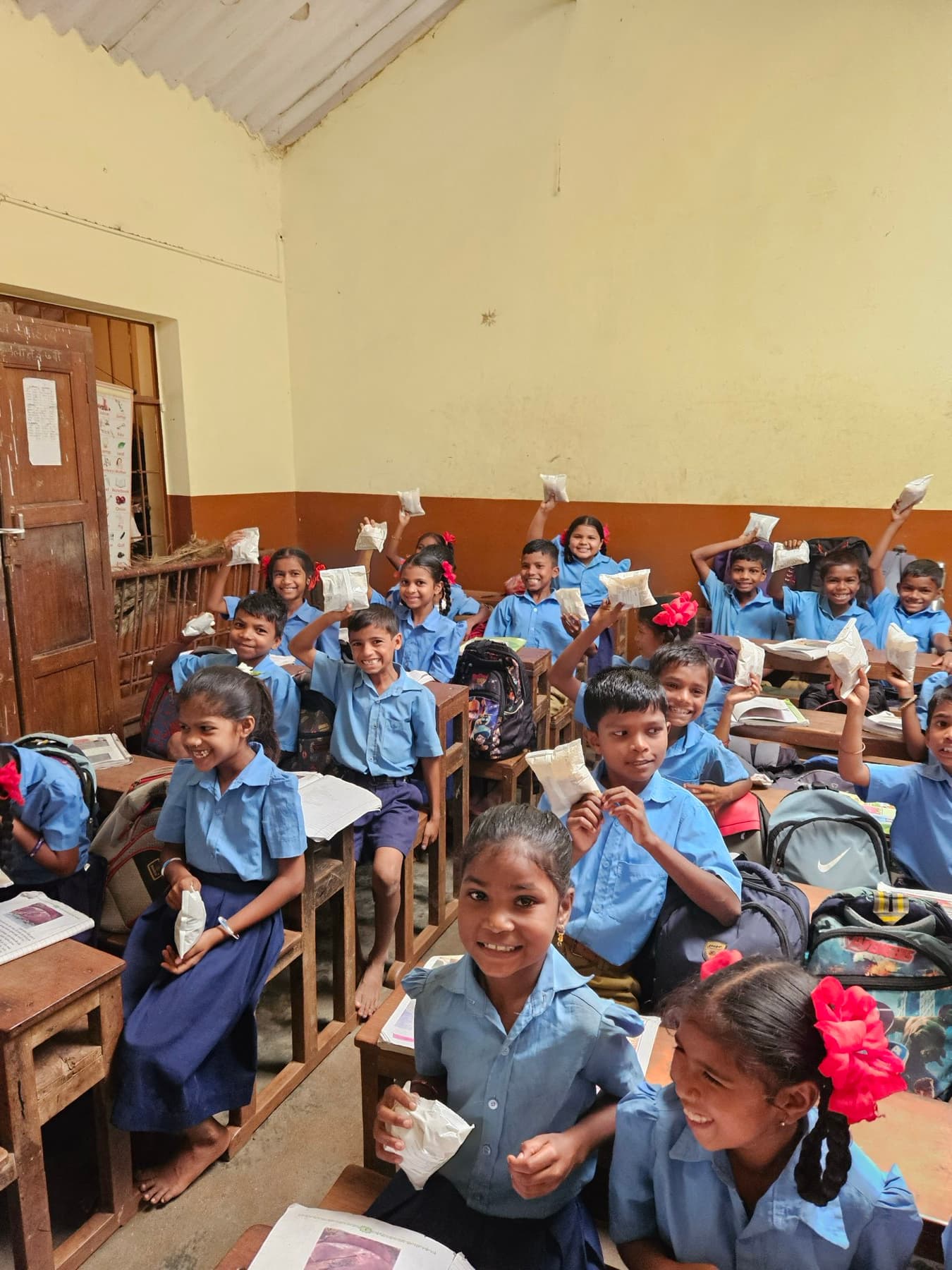 A full classroom of children hold up small nutrition packets together during a session.