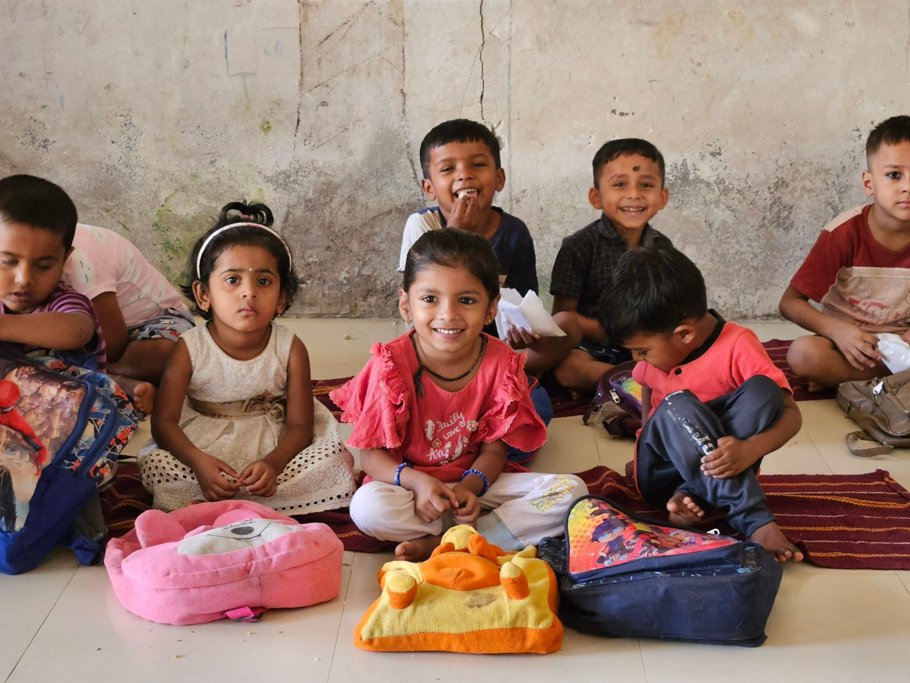 A group of schoolchildren sit together on benches opening their breakfast packets, one boy holding his up toward the camera.