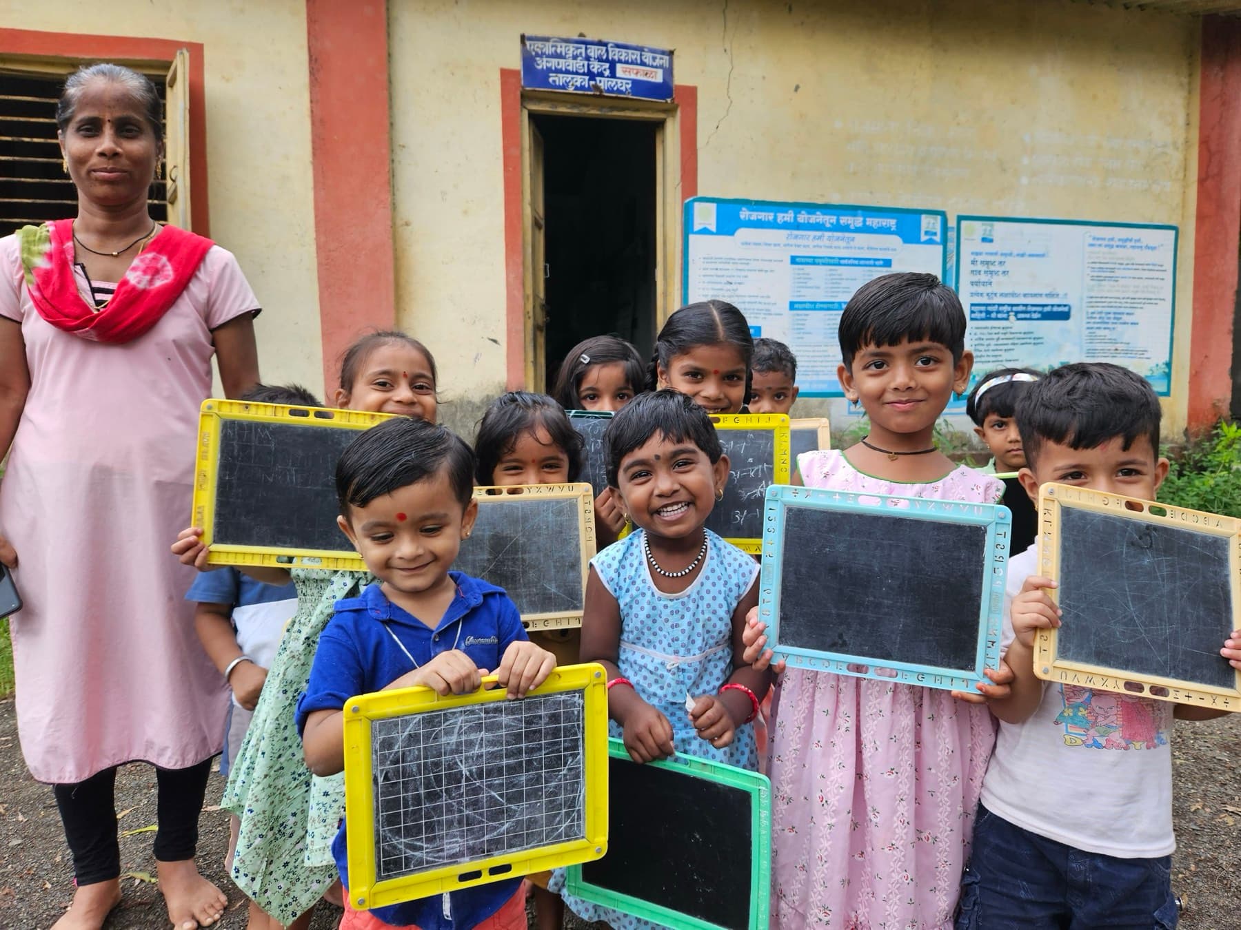 Anganwadi children hold up their writing slates beside their teacher.